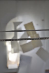 two trails of paper with words printed on, held taught in mid air, in front of a white wall which is covered in shadows.